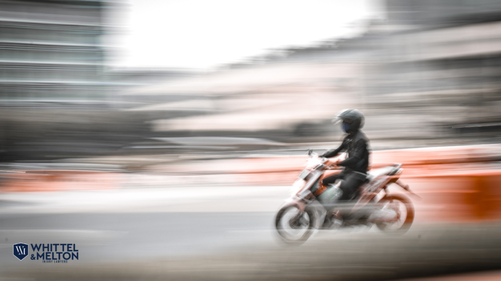 Whittel & Melton logo in the top left corner; a motorcyclist rides through a city street in Ocala, representing motorcycle accident in Ocala cases.