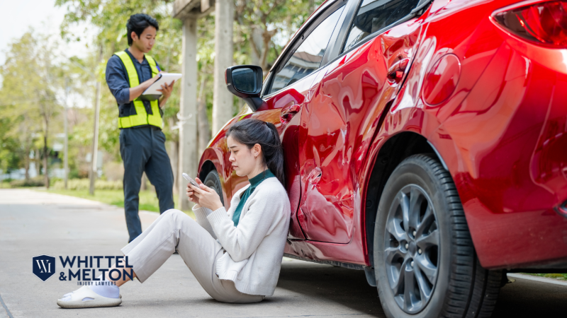 Woman sitting injured against a red car with visible side damage after an accident, checking her phone, while a man in a safety vest takes notes. Logo of Whittel & Melton leesburg car accident and injury attorney service.