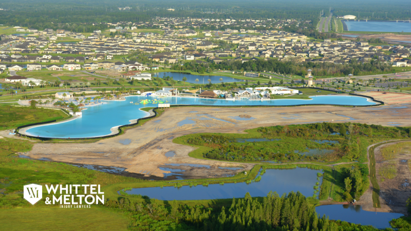 Aerial view of a large, bright blue man-made lagoon surrounded by new suburban housing in Pasco County, Florida, near US-19, with the logo for Whittel & Melton Injury Lawyers.